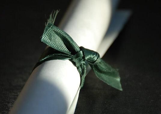 A rolled piece of white paper tied with green satin ribbon with light falling on the ribbon and the background being black.